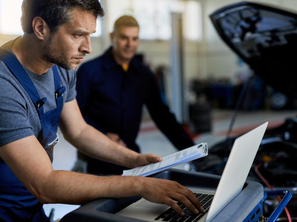 Auto repairman working on a computer while doing car diagnostic with his coworker in a workshop.